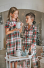 A young boy stands in a festively decorated room wearing Unisex Pj's in White Plaid. The classic 2 piece pajama set is buttery soft fabric.  The white background is accented by green, red and black plaid. There is red piping trim on the top. 