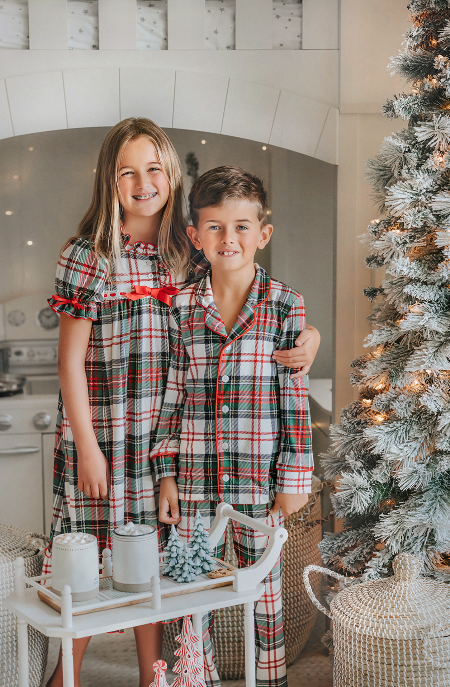 A young boy stands in a festively decorated room wearing Unisex Pj's in White Plaid. The classic 2 piece pajama set is buttery soft fabric.  The white background is accented by green, red and black plaid. There is red piping trim on the top. 