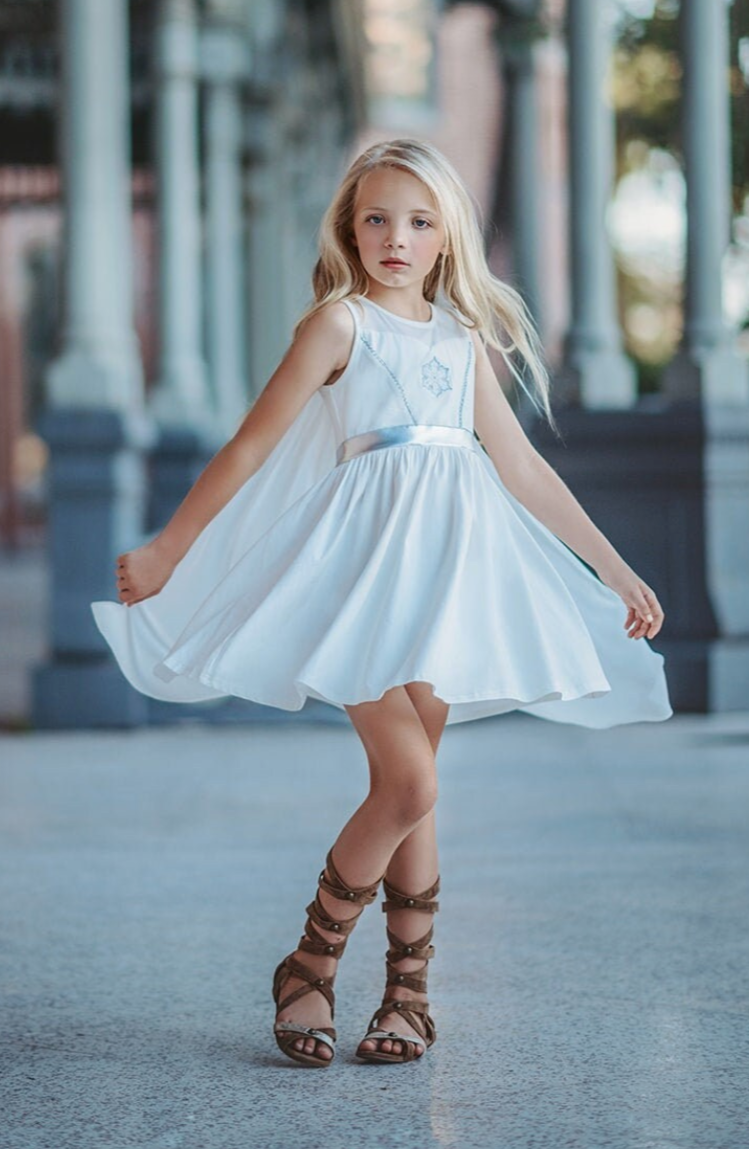 A young girl is wearing a white cotton twirl dress. The sleeveless dress has a snowflake embroidered in the center of the chest in silver. There is a silver band across the waist. A white cape is fastened to the back with buttons. 