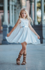 A young girl is wearing a white cotton twirl dress. The sleeveless dress has a snowflake embroidered in the center of the chest in silver. There is a silver band across the waist. A white cape is fastened to the back with buttons. 