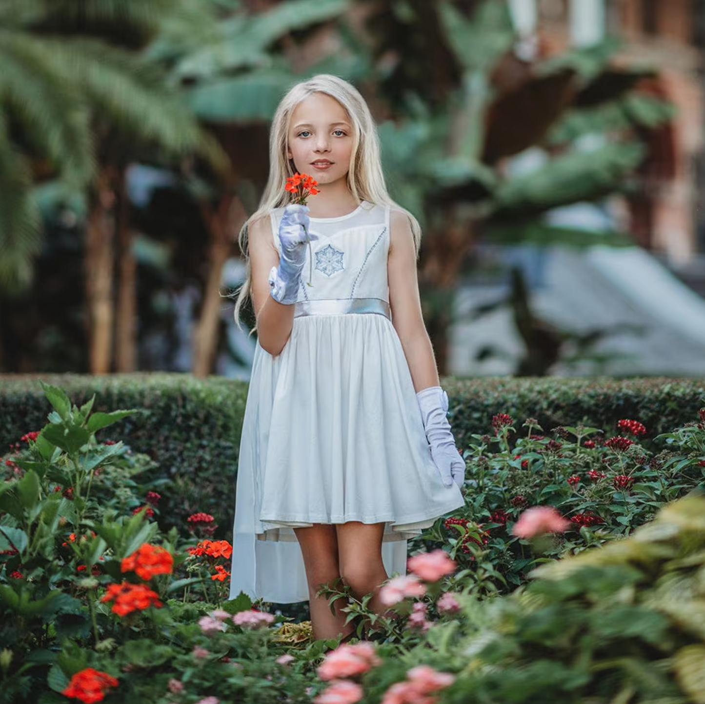 A young girl is wearing a white cotton twirl dress. The sleeveless dress has a snowflake embroidered in the center of the chest in silver. There is a silver band across the waist. A white cape is fastened to the back with buttons. 