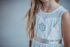 A young girl is wearing a white cotton twirl dress. The sleeveless dress has a snowflake embroidered in the center of the chest in silver. There is a silver band across the waist. A white cape is fastened to the back with buttons. 