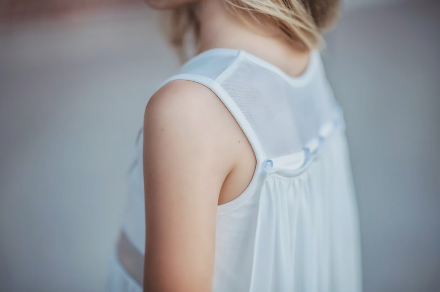 A young girl is wearing a white cotton twirl dress. The sleeveless dress has a snowflake embroidered in the center of the chest in silver. There is a silver band across the waist. A white cape is fastened to the back with buttons. 