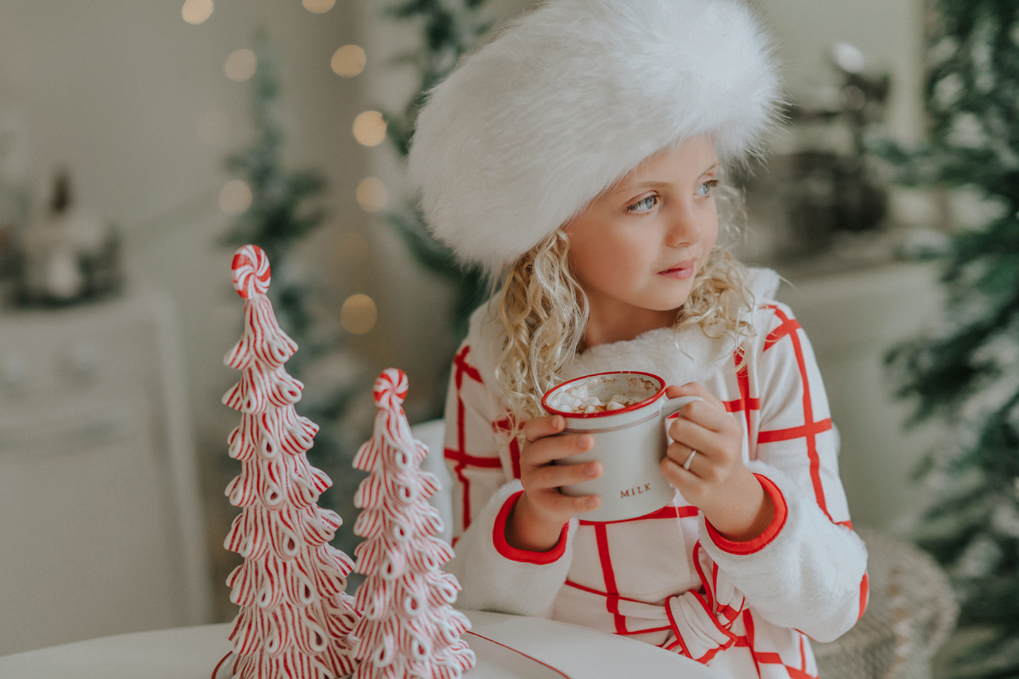 A young girl with curly blonde hair is wearing the Holiday Dancer- The Rockefeller Center Uniform Hat and a white coat with red plaid lines and red buttons. She is looking to the side, standing in a room with a softly blurred background and twinkling lights, embodying the charm of the Christmas Character Collection.