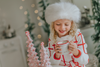 A young girl with curly blonde hair is wearing the Holiday Dancer- The Rockefeller Center Uniform Hat and a white coat with red plaid lines and red buttons. She is looking to the side, standing in a room with a softly blurred background and twinkling lights, embodying the charm of the Christmas Character Collection.