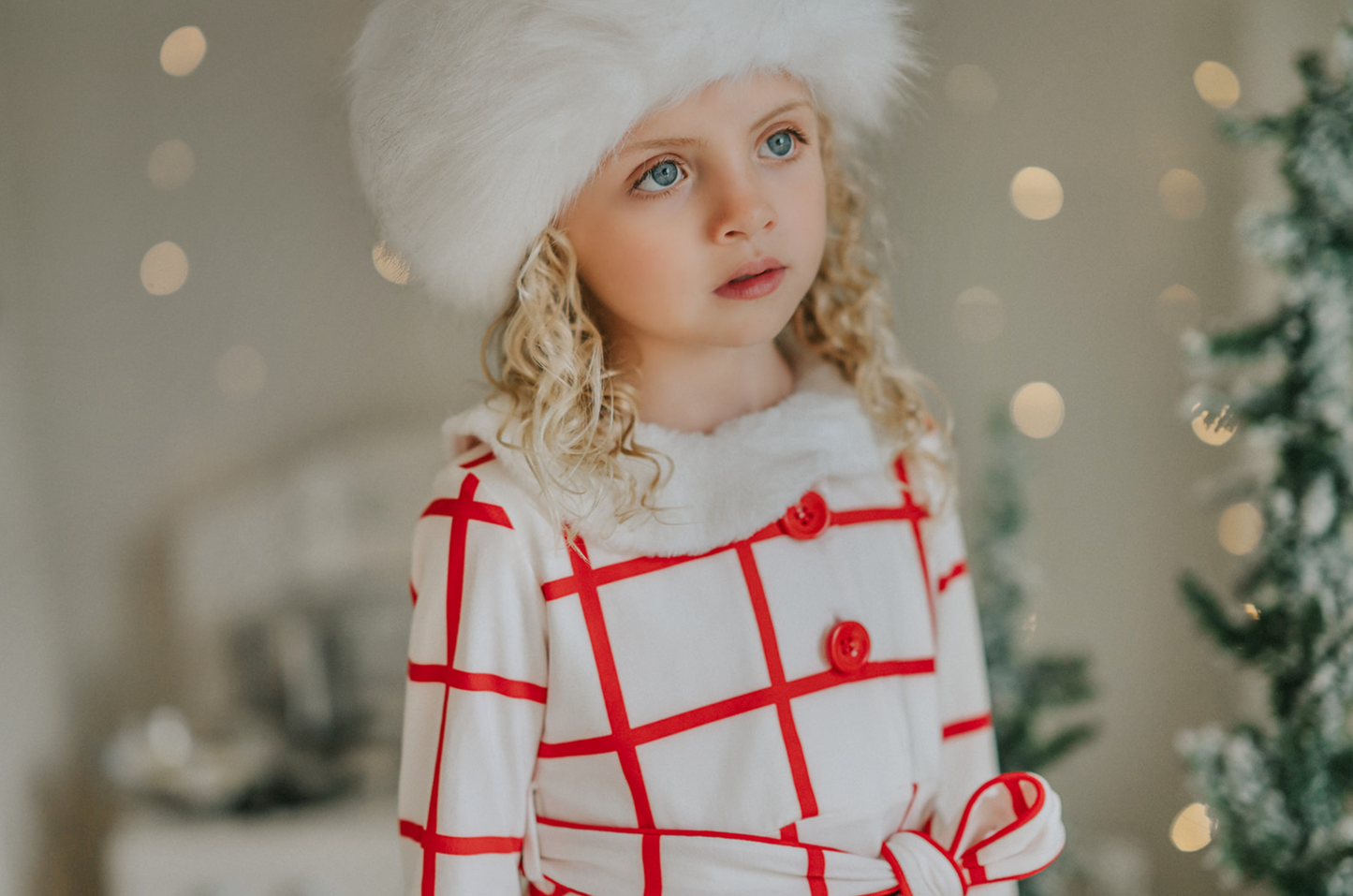 A young girl with curly blonde hair is wearing the Holiday Dancer- The Rockefeller Center Uniform Hat and a white coat with red plaid lines and red buttons. She is looking to the side, standing in a room with a softly blurred background and twinkling lights, embodying the charm of the Christmas Character Collection.