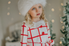 A young girl with curly blonde hair is wearing the Holiday Dancer- The Rockefeller Center Uniform Hat and a white coat with red plaid lines and red buttons. She is looking to the side, standing in a room with a softly blurred background and twinkling lights, embodying the charm of the Christmas Character Collection.