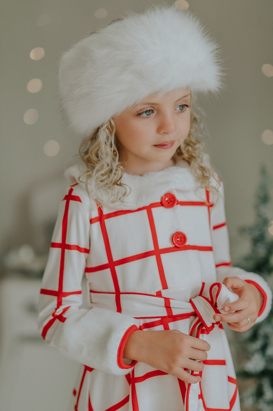 A young girl with curly blonde hair is wearing the Holiday Dancer- The Rockefeller Center Uniform Hat and a white coat with red plaid lines and red buttons. She is looking to the side, standing in a room with a softly blurred background and twinkling lights, embodying the charm of the Christmas Character Collection.