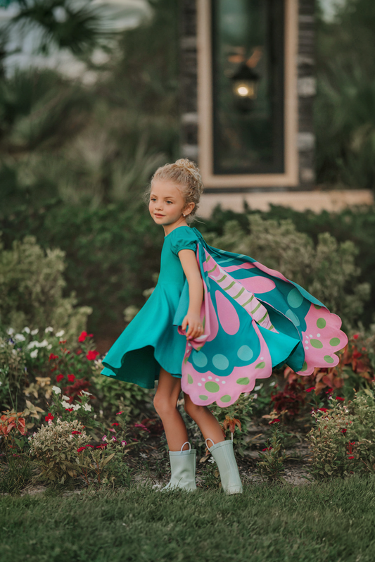 A young girl twirls in a beautiful turquoise dress.  The short puff sleeves and flowing skirt look classic.  There is a beautiful butterfly wing cape that attaches with buttons in the back.  The butterfly has bright teal, pink and green designs on the wings. 