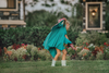 A young girl twirls in a beautiful turquoise dress.  The short puff sleeves and flowing skirt look classic.  There is a beautiful butterfly wing cape that attaches with buttons in the back.  The butterfly has bright teal, pink and green designs on the wings. 