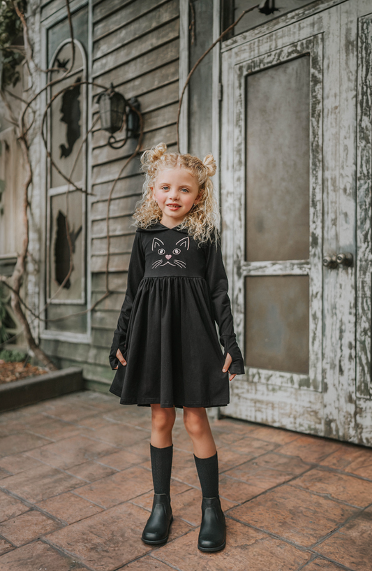 A young girl is wearing a black long sleeve cotton twirl dress.  There is a kitty embroidered on the chest.  The sleeves have pink kitty paws on them with thumbholes to put your thumb through.  Perfect for spooky season. 