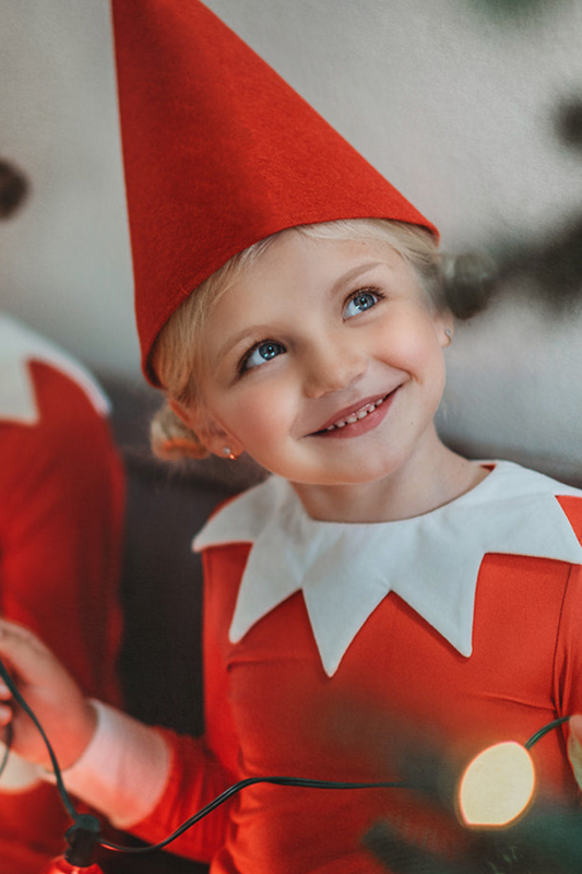 A young child wearing the Red Elf Hat, a red outfit with a white collar, and holding holiday lights, smiles while looking up. The background is blurred, emphasizing the child's joyful expression and festive fun, radiating Christmas cheer.