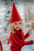 A young child wearing the Red Elf Hat, a red outfit with a white collar, and holding holiday lights, smiles while looking up. The hat is a cone shape and true red in color. The background is blurred, emphasizing the child's joyful expression and festive fun, radiating Christmas cheer.