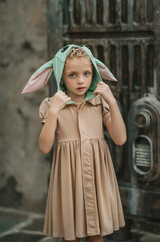 The Galactic Baby Alien Twirl Dress is a khaki color button up twirl dress.  The short puff sleeves are perfect for a hot park day.  The green hood is adorned with floppy ears lined with pink and large black eyes that remind you of your favorite baby alien that travels in a pod with a Mandolorian. 