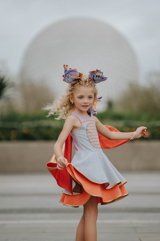 A young girl is wearing a light purple and heather grey cotton dress.  There is a light pink sparkle oval to resemble a belly.  The skirt is a double twirl with the second layer in orange.  The detachable wings are gold and orange. 