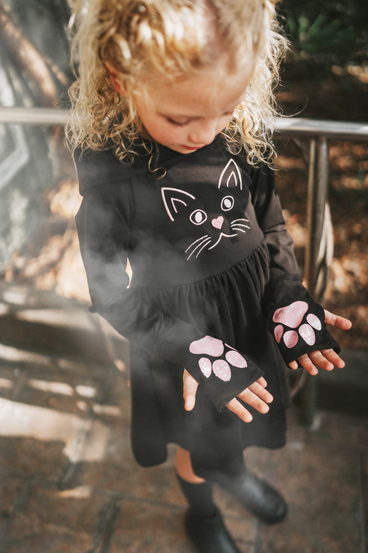 A young girl is wearing a black long sleeve cotton twirl dress.  There is a kitty embroidered on the chest.  The sleeves have pink kitty paws on them with thumbholes to put your thumb through.  Perfect for spooky season. 