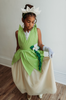 A young girl is wearing a beautiful bright green gown.  The layers on the bodice and skirt are cut like leaves.  There is a light yellow golden tulle skirt.   There is a magnolia flower with green leaves adorned on the side. 