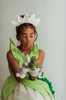 A young girl is wearing a beautiful bright green gown.  The layers on the bodice and skirt are cut like leaves.  There is a light yellow golden tulle skirt.   There is a magnolia flower with green leaves adorned on the side. 