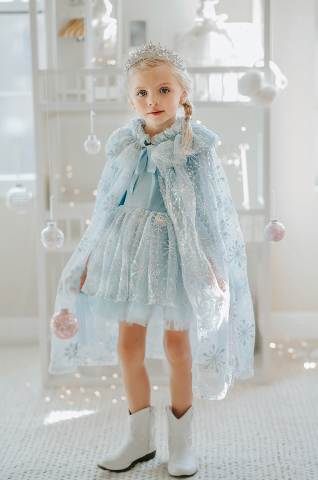 A young girl is wearing a light blue tutu dress. The velvet bodice has short sleeves.  The skirt has sparkling blue sequin accents shaped like snowflakes and falling snow.  There are light blue layers of tule giving a full tutu look. 