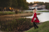 A young girl stands outdoors, wearing a vibrant red Pirate Twirl Dress with gold trim over a white ruffled shirt adorned with delicate ruffle detailing. She completes her outfit with black boots and a feathered black hat. The scenic background includes grassy areas, water, and trees, suggesting a park or natural setting.