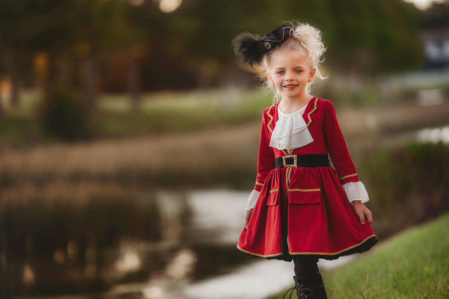 A young girl stands outdoors, wearing a vibrant red Pirate Twirl Dress with gold trim over a white ruffled shirt adorned with delicate ruffle detailing. She completes her outfit with black boots and a feathered black hat. The scenic background includes grassy areas, water, and trees, suggesting a park or natural setting.