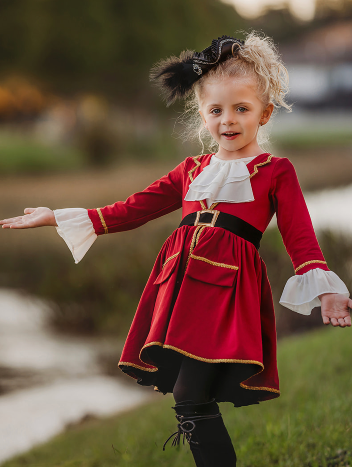 A young girl stands outdoors, wearing a vibrant red Pirate Twirl Dress with gold trim over a white ruffled shirt adorned with delicate ruffle detailing. She completes her outfit with black boots and a feathered black hat. The scenic background includes grassy areas, water, and trees, suggesting a park or natural setting.