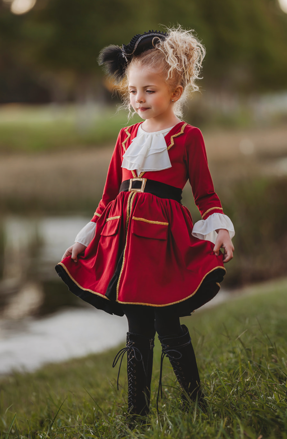 A young girl stands outdoors, wearing a vibrant red Pirate Twirl Dress with gold trim with a white ruffled neckline. Adorned with delicate ruffle detailing at the sleeves and a black waistband with gold buckle. 