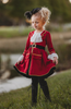 A young girl stands outdoors, wearing a vibrant red Pirate Twirl Dress with gold trim with a white ruffled neckline. Adorned with delicate ruffle detailing at the sleeves and a black waistband with gold buckle. 