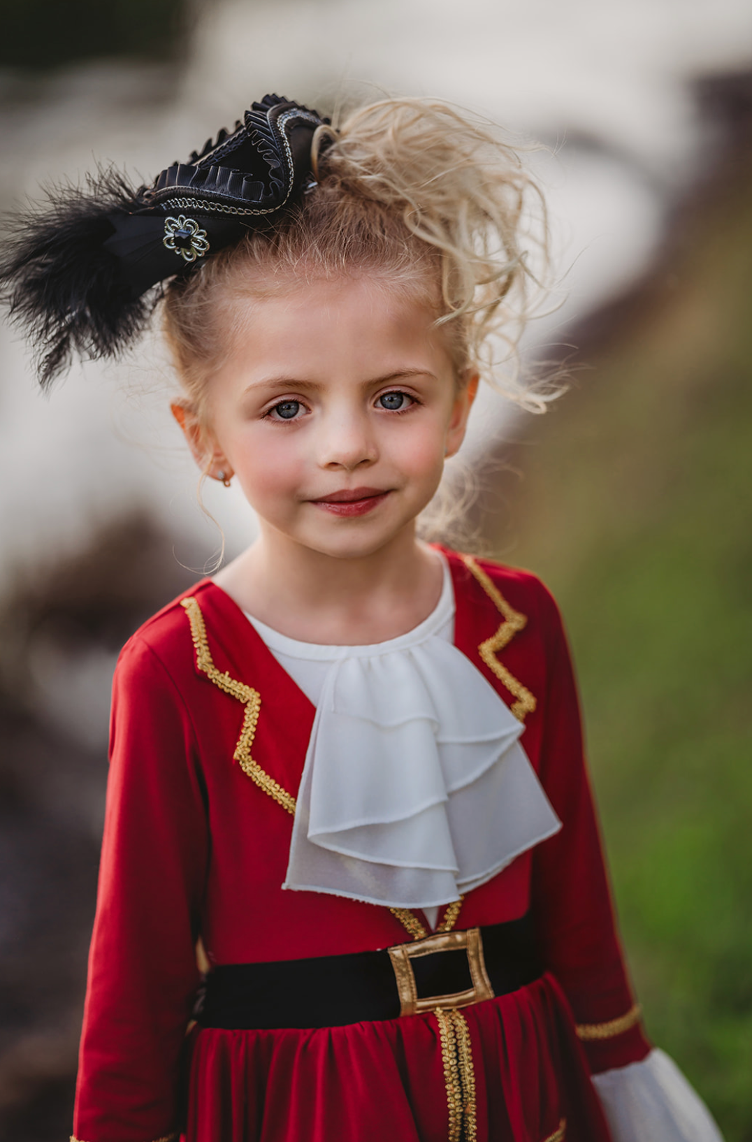 A young girl stands outdoors, wearing a vibrant red Pirate Twirl Dress with gold trim with a white ruffled neckline. Adorned with delicate ruffle detailing at the sleeves and a black waistband with gold buckle. 