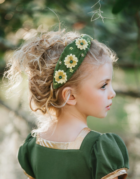 A young girl with curly blonde hair, wearing a green and gold maxi dress, is shown in profile. She is sporting the Flower Princess Headband, which is adorned with yellow and white flowers. The background features a blurred natural setting with greenery.