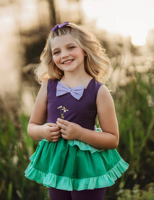 A young girl is wearing The Mermaid Peplum top.  The sleeveless top has bodice in deep purple.  There is a light purple bow at the neckline.  The peplum ruffle has a light green skirt over a green skirt with bottom ruffle.  There is tulle under the peplum and a cotton lining to add fullness. 