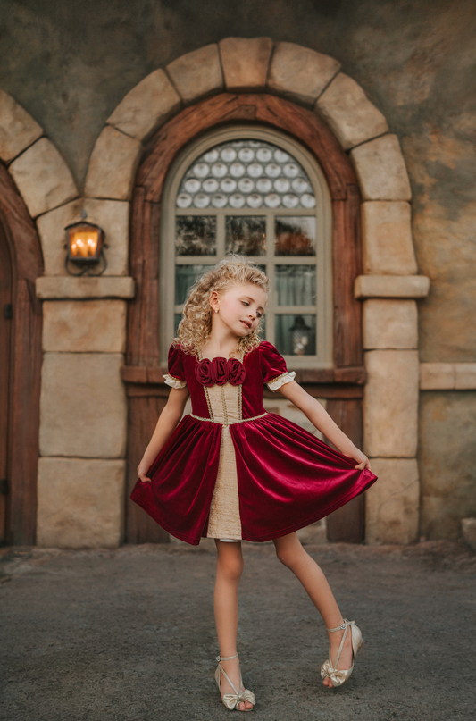 A young girl is wearing a twirl length gown inspired by our favorite red rose princess at Christmas.  The deep crimson red is stretch velvet.  The puff sleeves have a gold ruffle detail. Three chiffon roses line the neck and the bodice has gold inlay.  The skirt has gold inlay in a center pleat.  A stunning dress for any occasion. 