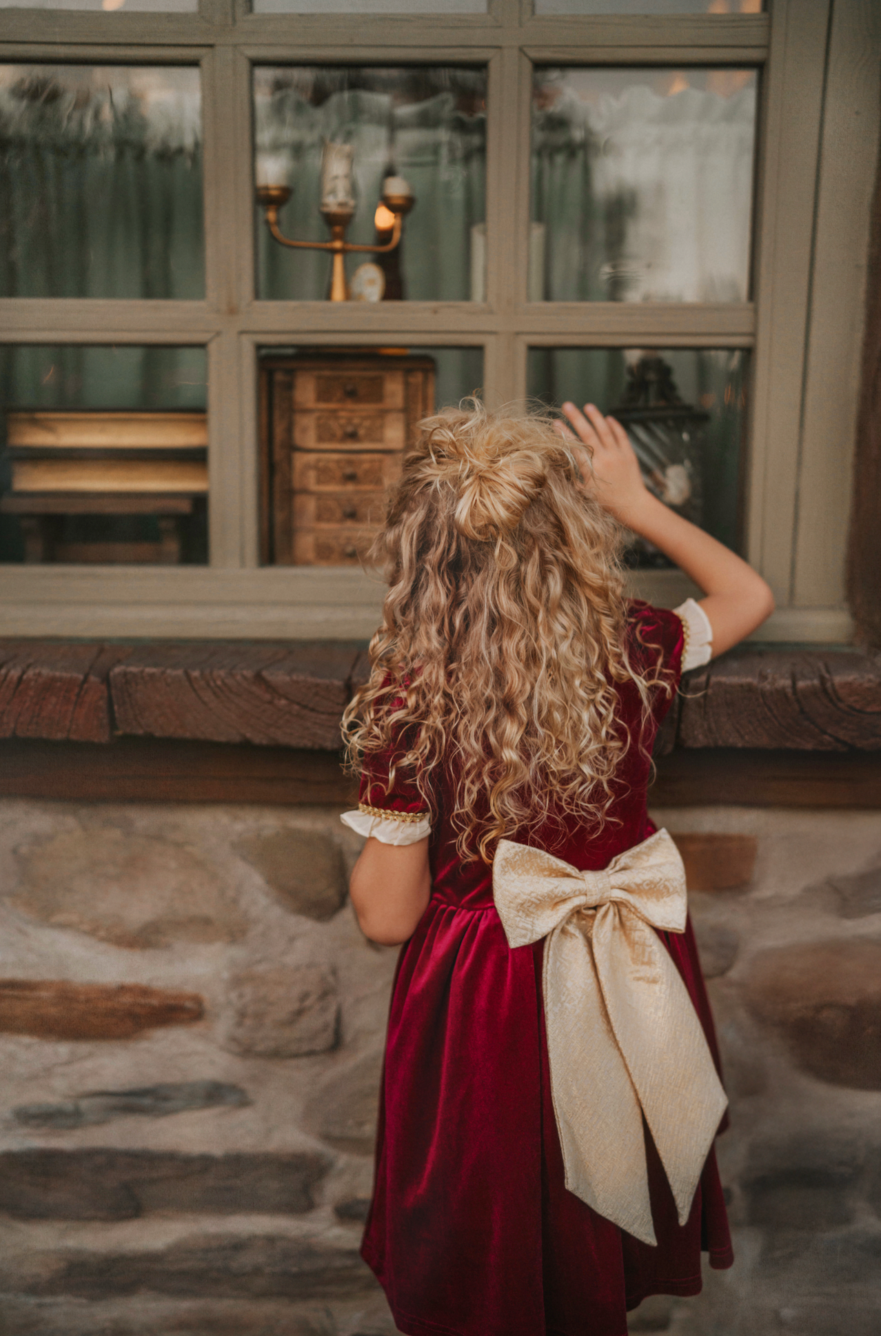 A young girl is wearing a twirl length gown inspired by our favorite red rose princess at Christmas.  The deep crimson red is stretch velvet.  The back has a large golden bow at the waist. The puff sleeves have a gold ruffle detail. Three chiffon roses line the neck and the bodice has gold inlay.  The skirt has gold inlay in a center pleat.  A stunning dress for any occasion. 