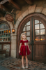 A young girl is wearing a twirl length gown inspired by our favorite red rose princess at Christmas.  The deep crimson red is stretch velvet.  The puff sleeves have a gold ruffle detail. Three chiffon roses line the neck and the bodice has gold inlay.  The skirt has gold inlay in a center pleat.  A stunning dress for any occasion. 