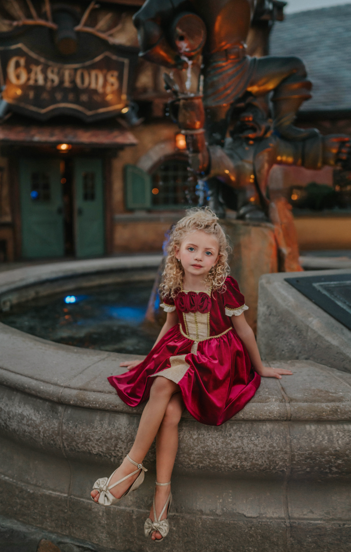 A young girl is wearing a twirl length gown inspired by our favorite red rose princess at Christmas.  The deep crimson red is stretch velvet.  The puff sleeves have a gold ruffle detail. Three chiffon roses line the neck and the bodice has gold inlay.  The skirt has gold inlay in a center pleat.  A stunning dress for any occasion. 