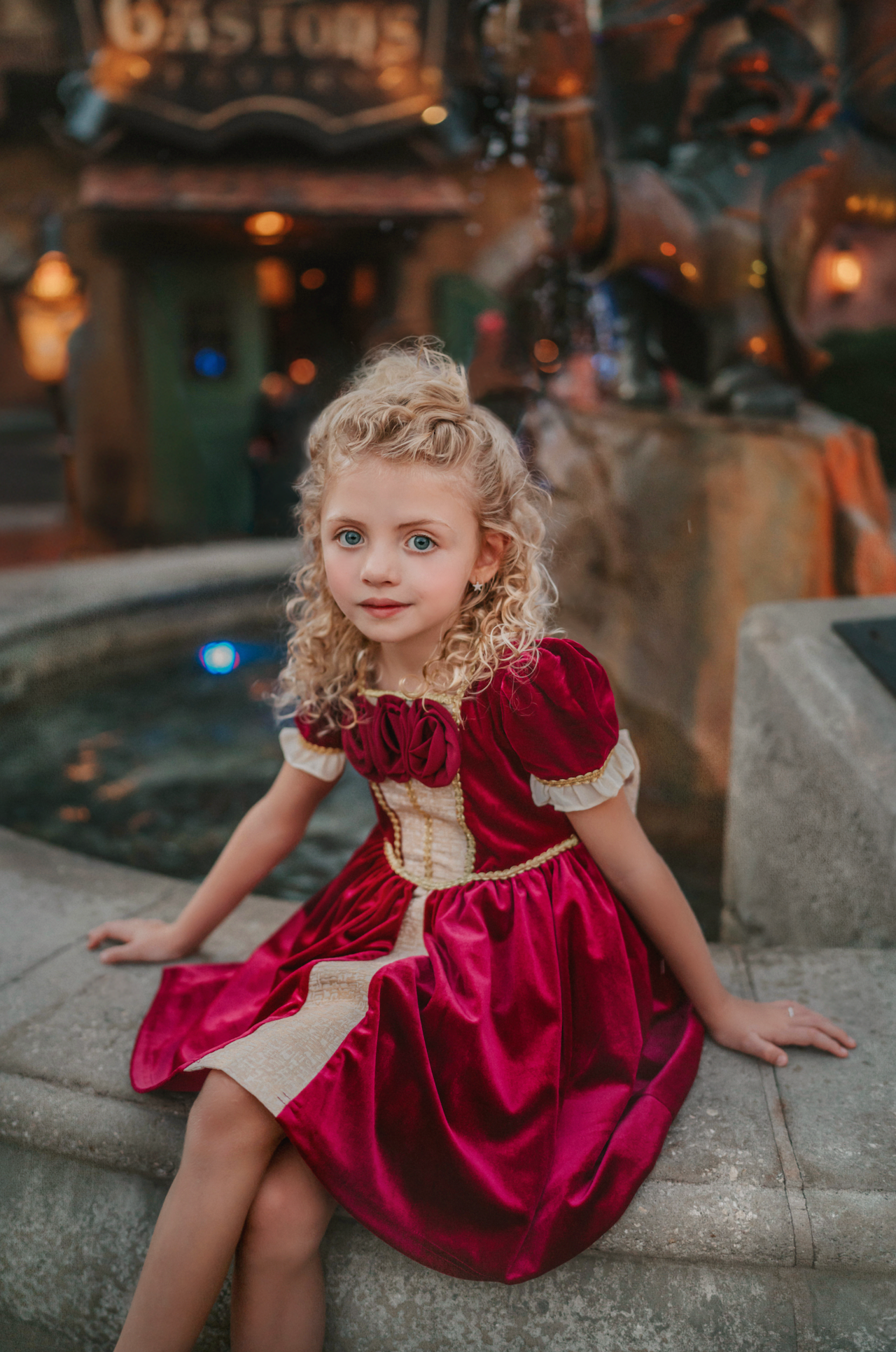 A young girl is wearing a twirl length gown inspired by our favorite red rose princess at Christmas.  The deep crimson red is stretch velvet.  The puff sleeves have a gold ruffle detail. Three chiffon roses line the neck and the bodice has gold inlay.  The skirt has gold inlay in a center pleat.  A stunning dress for any occasion. 