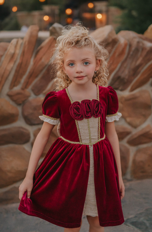 A young girl is wearing a twirl length gown inspired by our favorite red rose princess at Christmas.  The deep crimson red is stretch velvet.  The puff sleeves have a gold ruffle detail. Three chiffon roses line the neck and the bodice has gold inlay.  The skirt has gold inlay in a center pleat.  A stunning dress for any occasion. 