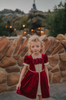 A young girl is wearing a twirl length gown inspired by our favorite red rose princess at Christmas.  The deep crimson red is stretch velvet.  The puff sleeves have a gold ruffle detail. Three chiffon roses line the neck and the bodice has gold inlay.  The skirt has gold inlay in a center pleat.  A stunning dress for any occasion. 