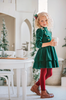 A young girl is wearing an evergreen velvet soldier twirl dress.  The deep velvet is stretchy and has a uniform collar with gold buttons down the bodice.  The 3/4 sleeves have elastic at the cuff.  There are two layers of velvet in the skirts. 