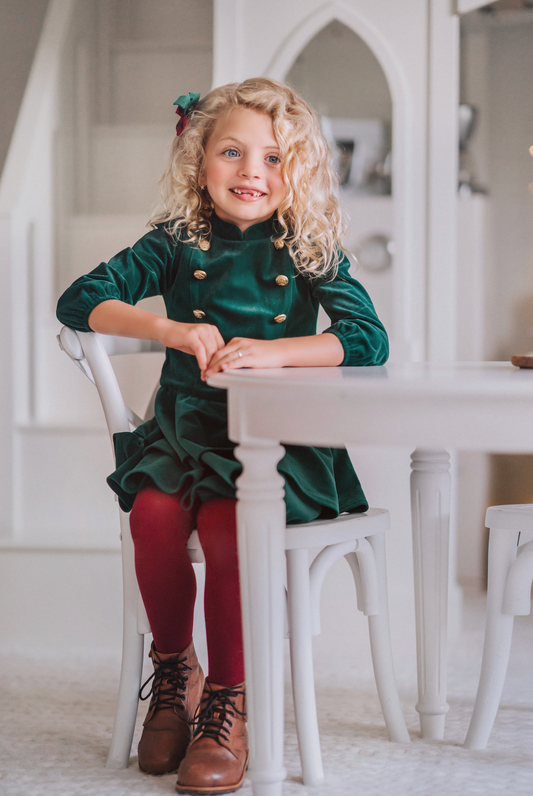 A young girl is wearing an evergreen velvet soldier twirl dress.  The deep velvet is stretchy and has a uniform collar with gold buttons down the bodice.  The 3/4 sleeves have elastic at the cuff.  There are two layers of velvet in the skirts. 