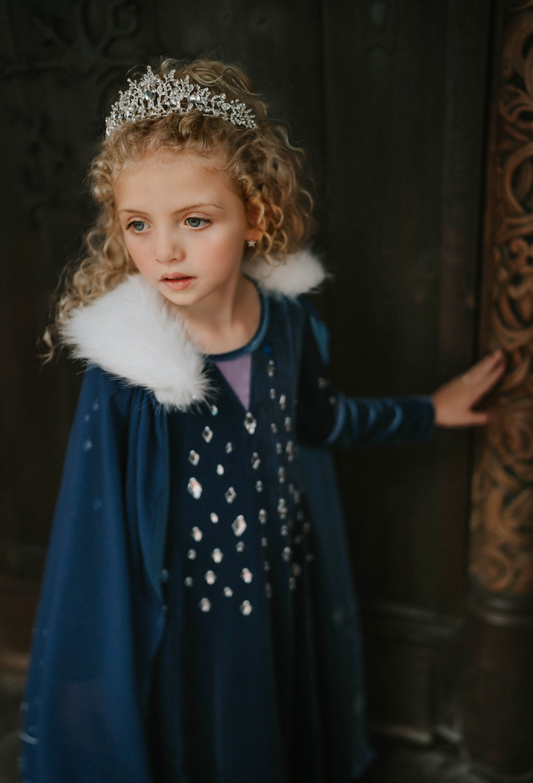 A young girl is wearing a navy blue chiffon cape with white faux fur trim.  The matching velvet gown is adorned with diamond shape crystal gems of various sizes.  There is a small lavender triangle near the neck. 