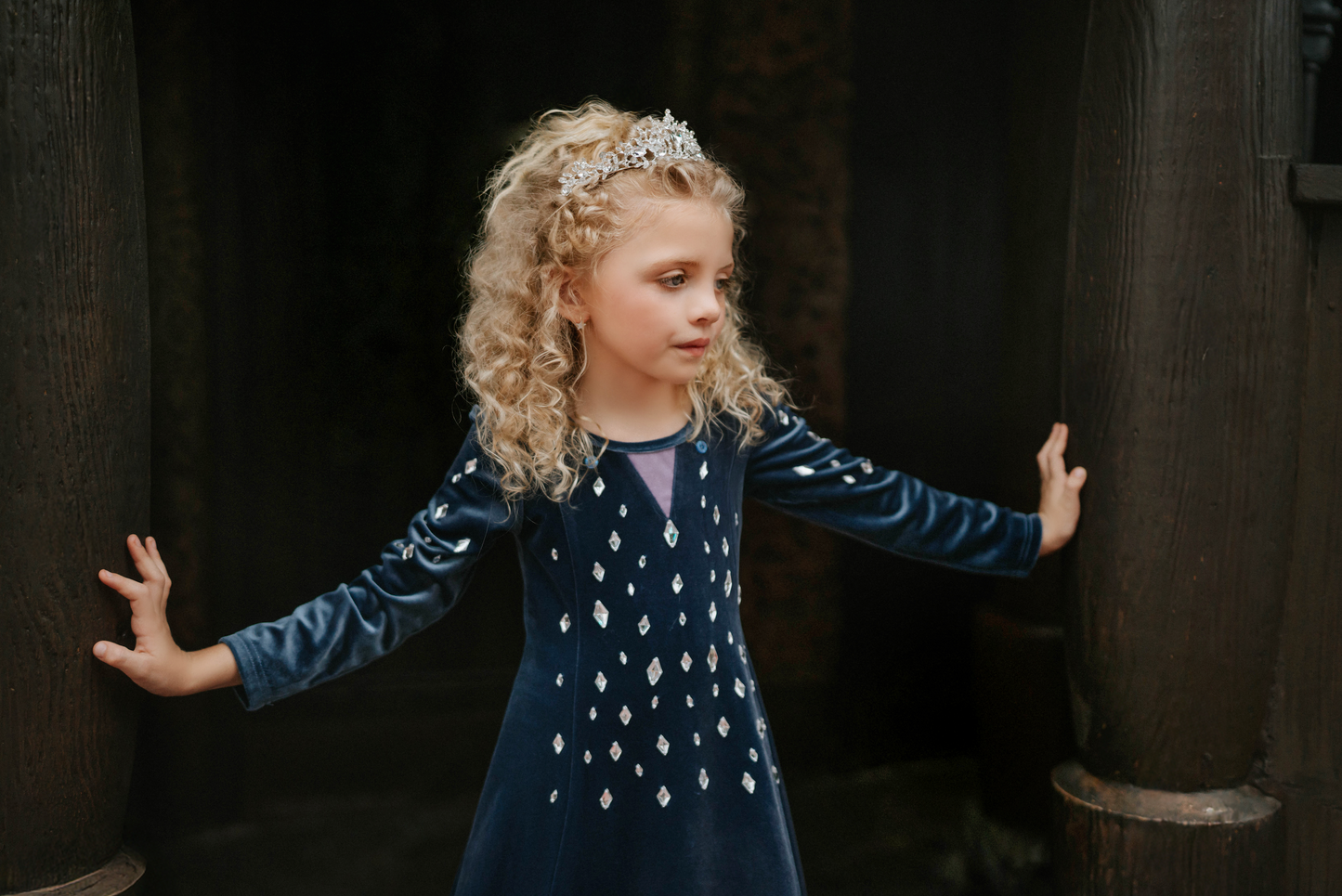 A young girl is wearing a navy blue velvet gown is adorned with diamond shape crystal gems of various sizes.  There is a small lavender triangle near the neck. 