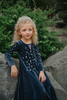 A young girl is wearing a navy blue velvet gown is adorned with diamond shape crystal gems of various sizes.  There is a small lavender triangle near the neck. 