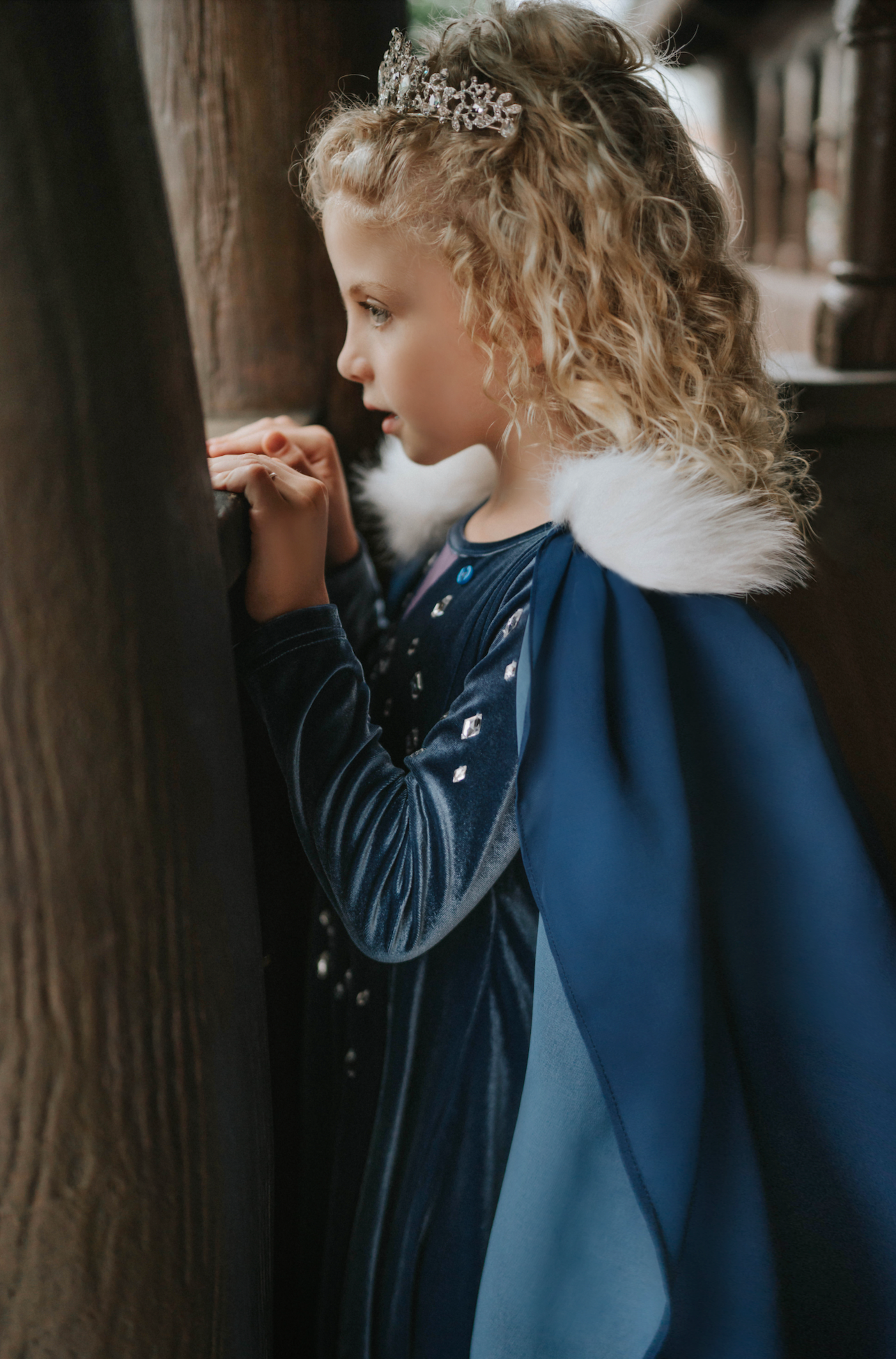 A young girl is wearing a navy blue velvet gown is adorned with diamond shape crystal gems of various sizes.  There is a small lavender triangle near the neck. She has on a navy blue chiffon cape with white faux fur trim on the collar.