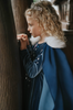 A young girl is wearing a navy blue velvet gown is adorned with diamond shape crystal gems of various sizes.  There is a small lavender triangle near the neck. She has on a navy blue chiffon cape with white faux fur trim on the collar.