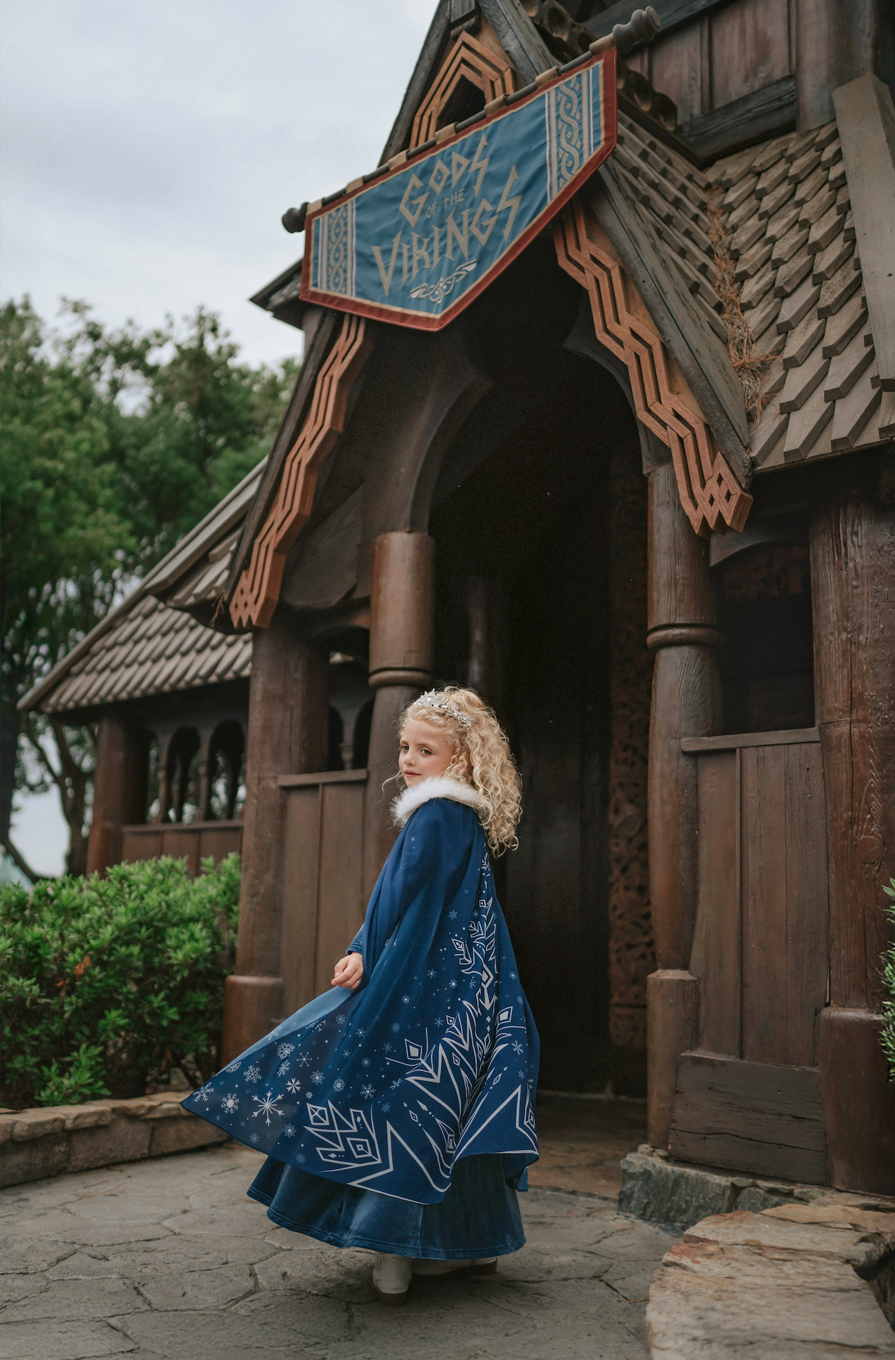 A young girl is wearing a navy blue velvet gown is adorned with diamond shape crystal gems of various sizes.  There is a small lavender triangle near the neck. She has on a navy blue chiffon cape with white faux fur trim on the collar. There is a snow flake design on the back of the cape.