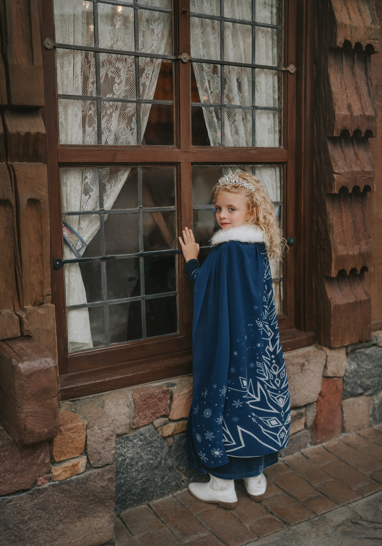 A young girl is wearing a navy blue velvet gown is adorned with diamond shape crystal gems of various sizes.  There is a small lavender triangle near the neck. She has on a navy blue chiffon cape with white faux fur trim on the collar. There is a snow flake design on the back of the cape.