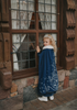 A young girl is wearing a navy blue velvet gown is adorned with diamond shape crystal gems of various sizes.  There is a small lavender triangle near the neck. She has on a navy blue chiffon cape with white faux fur trim on the collar. There is a snow flake design on the back of the cape.