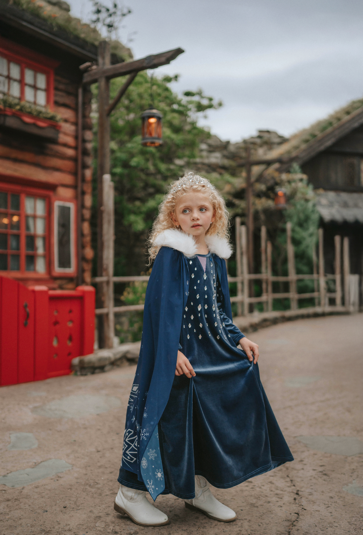 A young girl is wearing a navy blue velvet gown is adorned with diamond shape crystal gems of various sizes.  There is a small lavender triangle near the neck. She has on a navy blue chiffon cape with white faux fur trim on the collar. There is a snow flake design on the back of the cape.