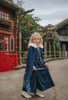 A young girl is wearing a navy blue velvet gown is adorned with diamond shape crystal gems of various sizes.  There is a small lavender triangle near the neck. She has on a navy blue chiffon cape with white faux fur trim on the collar. There is a snow flake design on the back of the cape.